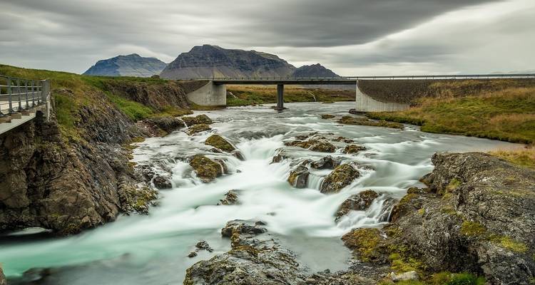 Rushing river under a bridge with mountainous backdrop.