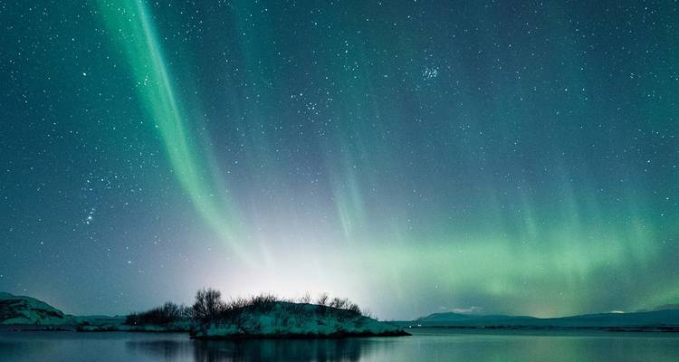 Des arcs vibrants d'aurores boréales s'étirent au-dessus d'un lac islandais tranquille et d'une île couverte de neige sous des cieux étoilés.