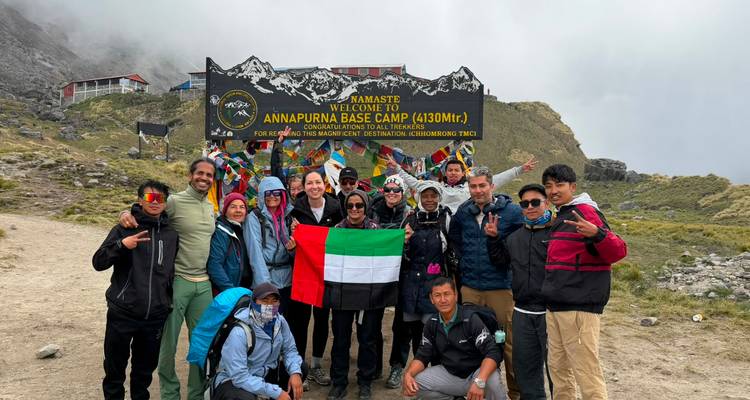Large trekking group poses with national flag in front of the Annapurna Base Camp welcome sign on a cloudy day.