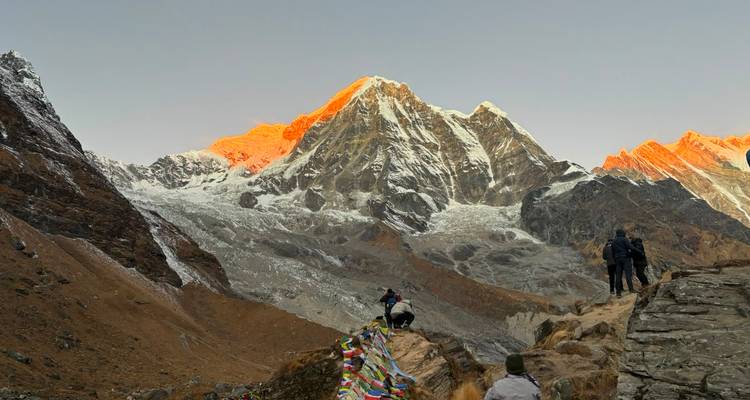 Hikers with prayer flags admire Himalayan peaks glowing orange at daybreak.