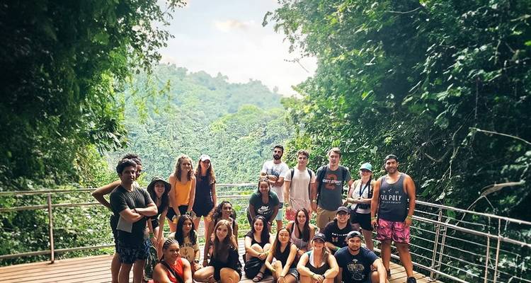 Grupo grande de viajeros posando en una cubierta con vista a densas colinas de selva tropical.