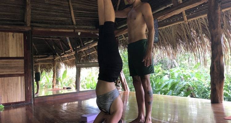 Woman doing a headstand with assistance in a thatched roof structure.