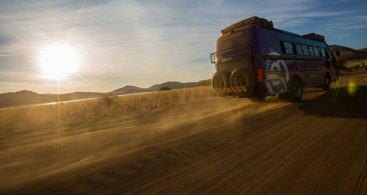 El autobús púrpura de expedición terrestre levanta polvo en el camino mientras el amanecer ilumina la carretera abierta de la sabana