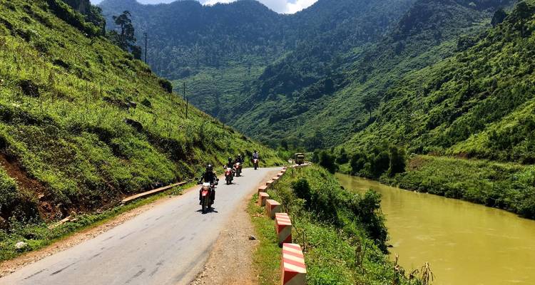 People riding motorcycles on a scenic road by a river in a mountainous area.