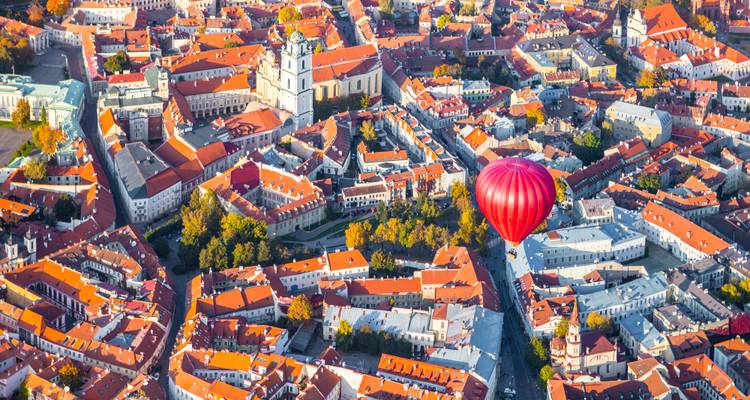 Toma aérea espectacular del Casco Antiguo de Vilnius con sus edificios de techos rojos enmarcados por árboles otoñales y un único globo aerostático rojo brillante flotando arriba bajo una cálida luz dorada.