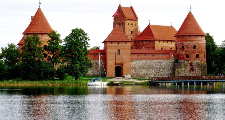 El Castillo de la Isla de Trakai de color rojo ladrillo reflejado en un lago tranquilo rodeado de vegetación bajo un cielo pálido y nublado.