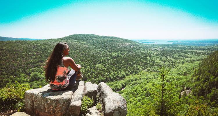 Randonneuse assise sur un rebord rocheux surplombant des vallées boisées luxuriantes et de l'eau au loin en Acadie.