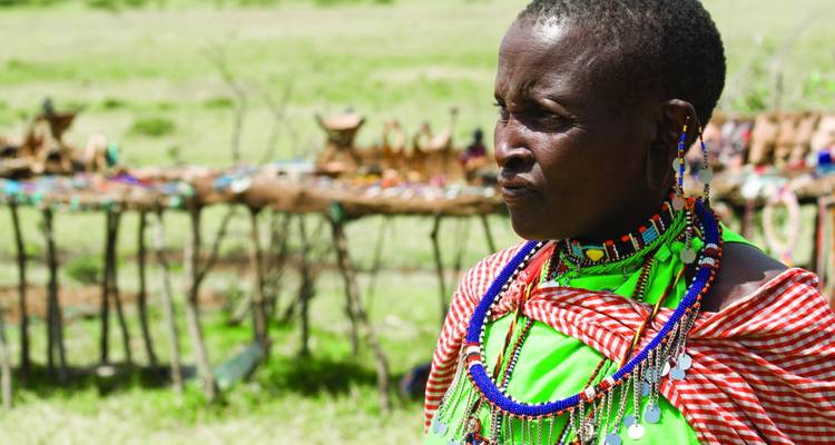 Retrato de una mujer maasai vistiendo ropa verde vibrante y joyería elaborada con cuentas intrincadas con un puesto de artesanías desenfocado detrás de ella.