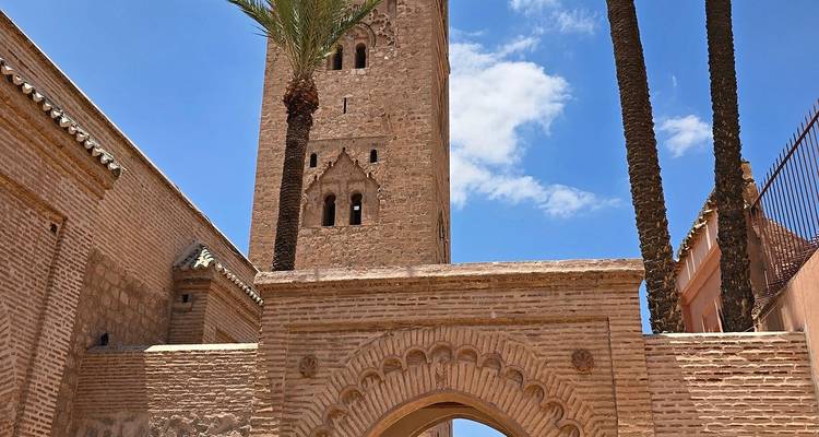 Minarete de piedra de una mezquita histórica se eleva detrás de una entrada arqueada de ladrillo enmarcada por altas palmeras bajo un cielo azul brillante.