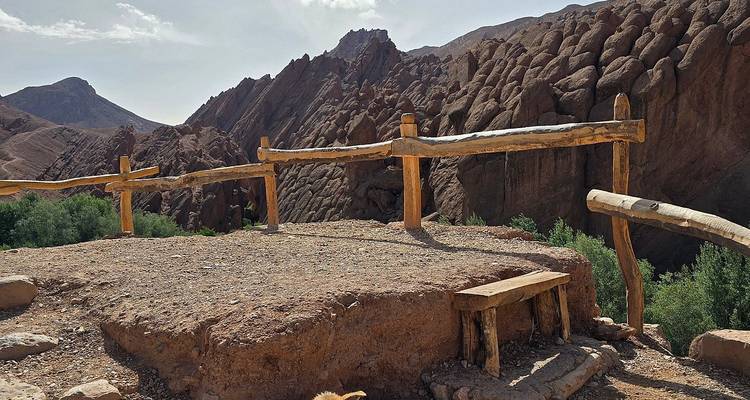 Mirador de cañón desértico rocoso con barandilla de madera y un banco sencillo con vista a acantilados rojos escarpados y vegetación escasa.