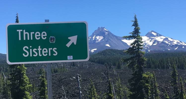 Sign pointing to Three Sisters mountains with scenic backdrop.