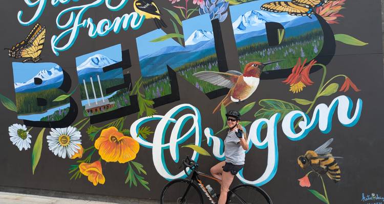 A person posing with a mural reading 'Greetings from Bend Oregon'.