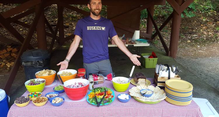 Man presenting a variety of food on a covered picnic table.