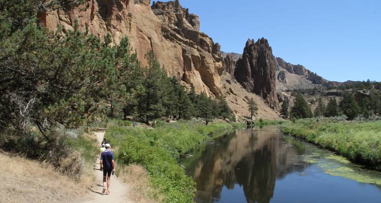 Personne marchant sur un sentier au bord d'une rivière, entourée d'arbres et de falaises rocheuses.