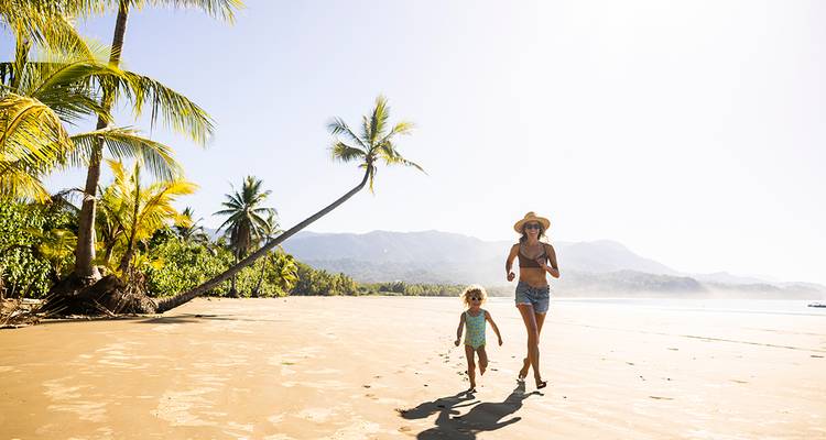 Mère et enfant courant le long d'une belle plage avec des palmiers and des montagnes en arrière-plan.