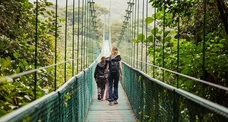 Deux personnes marchant sur un pont suspendu dans une forêt tropicale luxuriante, possiblement à Monteverde.