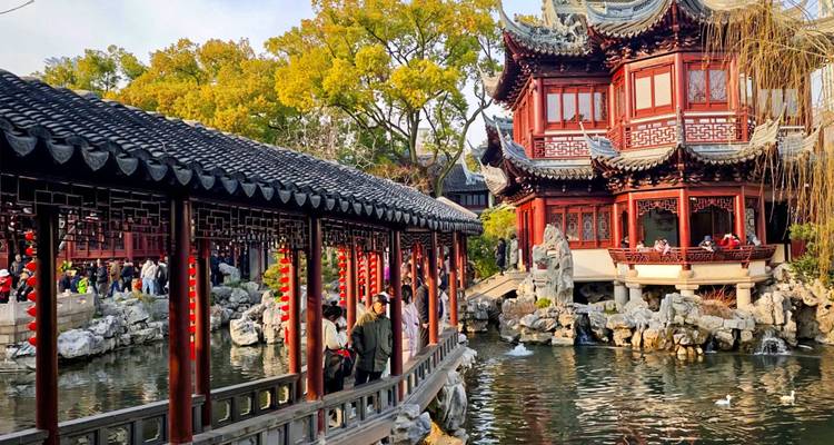 Allée couverte traditionnelle et pavillon orné à côté d'un bassin réfléchissant dans le jardin Yuyuan avec des visiteurs se promenant et des arbres d'automne en arrière-plan.