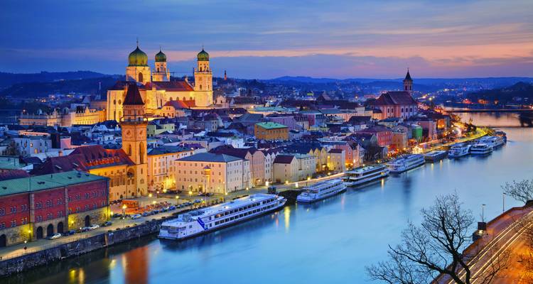Twilight panorama of Passau’s old town at the confluence of rivers with brightly lit cathedral domes and cruise boats reflecting off calm waters.