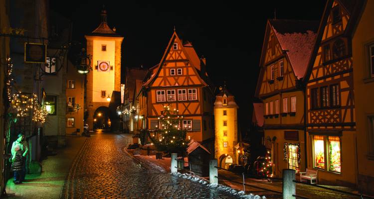 Snow-dusted cobblestone street in Rothenburg ob der Tauber lined with half-timbered houses and twinkling holiday lights at night.