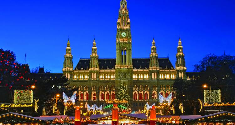 Vienna City Hall illuminated above a festive Christmas market with sparkling lights, angels and a towering tree against a deep blue twilight sky.