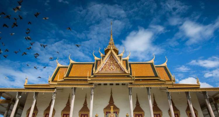 Toit doré orné et flèches du Palais Royal de Phnom Penh se détachant sur un ciel bleu éclatant avec des pigeons en vol.