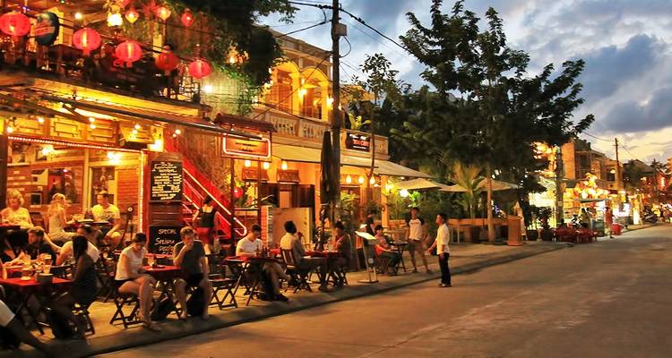 Calle animada con gente cenando al aire libre por la noche.