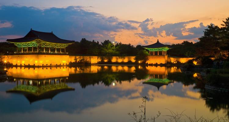 Pabellón coreano tradicional iluminado en oro que se refleja perfectamente en un estanque tranquilo al atardecer.