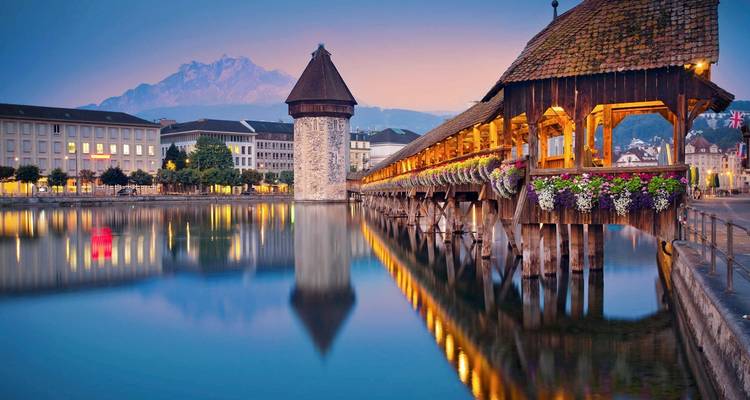Chapel Bridge over calm water at sunset with mountains in the background.