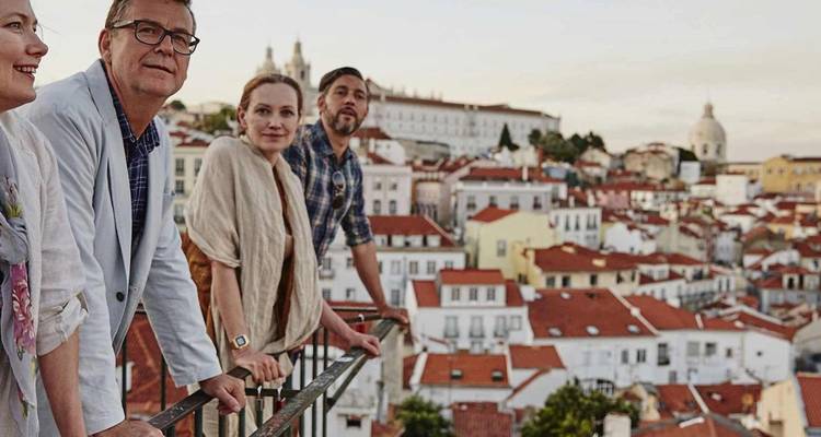 Cuatro adultos se apoyan en la barandilla de un mirador con vista al barrio de Alfama de techos rojos en Lisboa al anochecer.