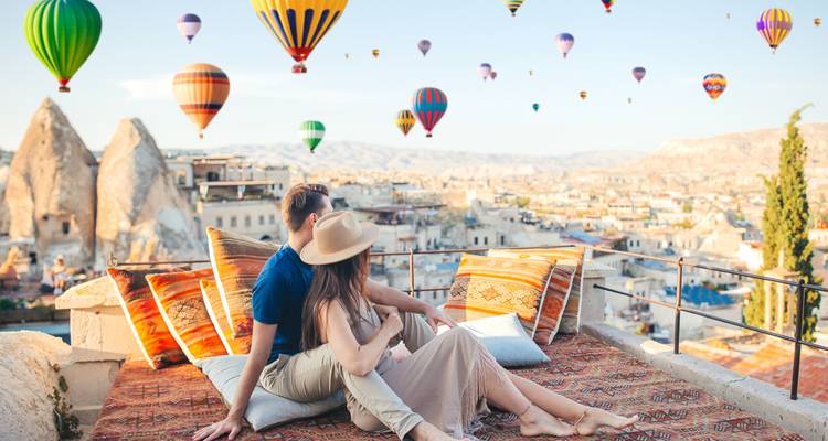 Romantisch koppel ontspant op een dakterras terwijl kleurrijke luchtballonnen over de rotsachtige landschappen van Cappadocië drijven bij dageraad.