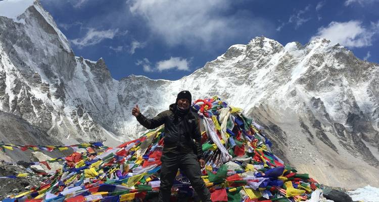 A person standing in front of colorful prayer flags with snowy mountains behind.