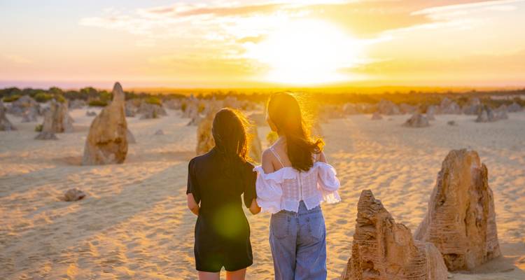 Dos mujeres jóvenes pasean por el dorado Desierto de los Pináculos al atardecer con torres de arenisca a su alrededor.