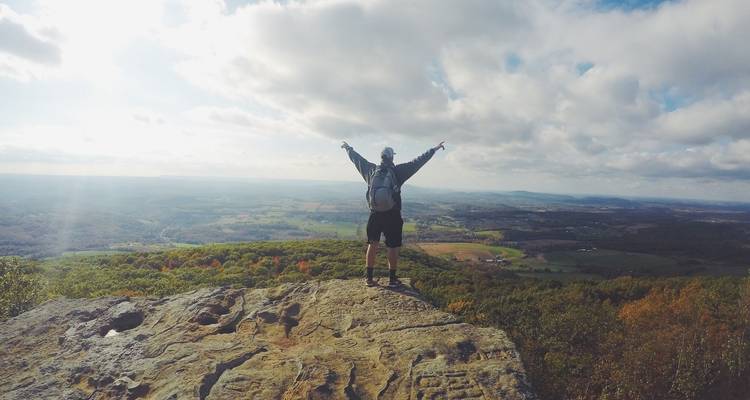 Eine Person, die triumphierend auf einer felsigen Klippe steht und weite Landschaften überblickt.