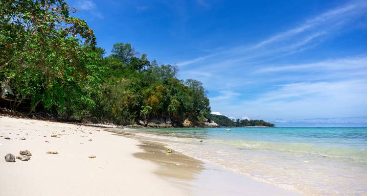 Ongerepte tropische strand met wit zand, turquoise water en weelderige met bomen bedekte kaap onder blauwe hemel.