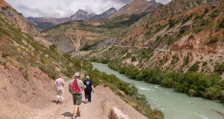 Los excursionistas siguen un sendero junto al río a través de un dramático cañón de laderas rojas y verdes hacia picos de Asia Central cubiertos de nieve