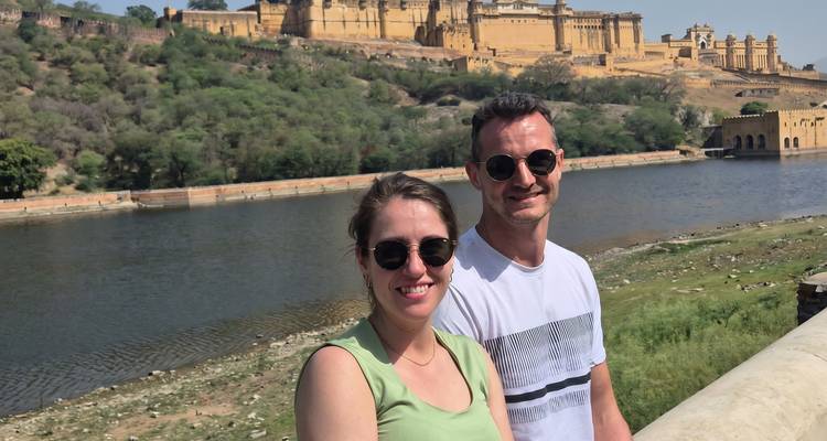 Smiling couple posing by a riverside with the yellow sandstone Amer Fort rising on the hill behind them