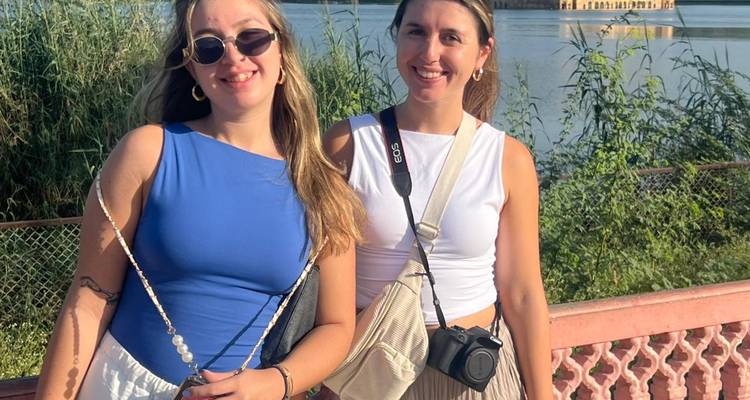 Two young women smiling by a railing with lake and Jal Mahal palace in the background
