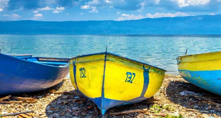 Des petits bateaux de pêche aux couleurs vives tirés sur un rivage de lac pavé de galets, avec des montagnes turquoise de l'autre côté de l'eau.
