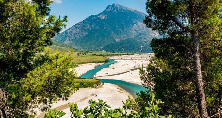 Un río turquesa serpenteante corta a través de un amplio valle arenoso bajo un pico imponente.