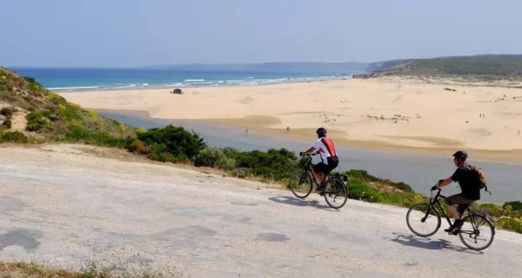 Twee fietsers rijden langs een kustweg met uitzicht op een breed zandstrand en blauwe oceaanbaai onder een wolkenloze hemel