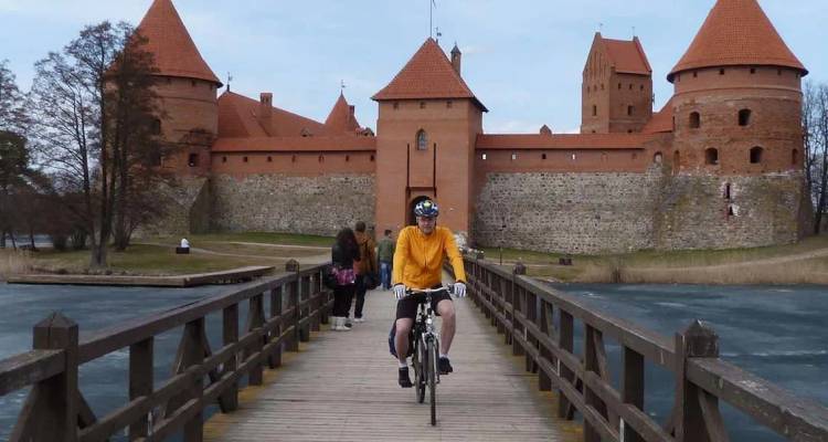 Cyclist rides across a wooden bridge toward the red-brick towers of Trakai Castle beside a frozen moat.