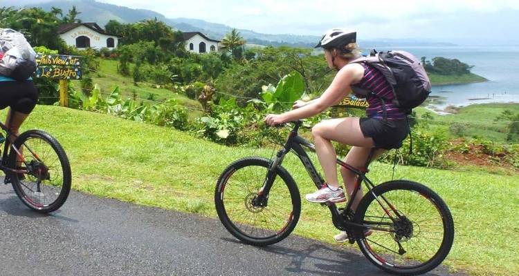 Dos ciclistas andan en bicicletas de montaña por un camino a orilla del lago rodeado de una exuberante naturaleza tropical en Costa Rica.