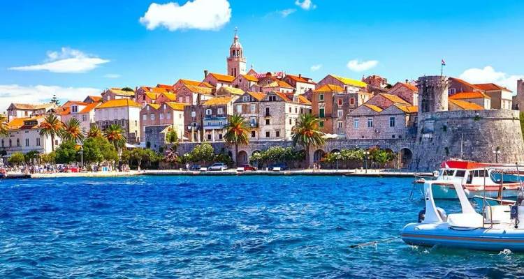 Pintoresco paseo marítimo medieval de Korčula con tejados naranjas y una torre de piedra frente al azul vibrante del Adriático.