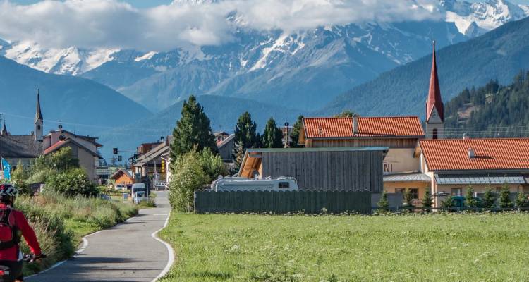 Un ciclista recorre un sendero pastoral hacia pueblos alpinos rodeados de imponentes picos nevados.