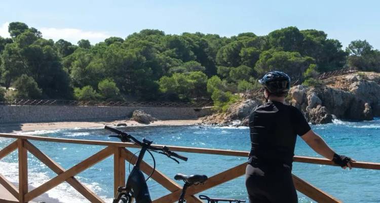 Cycliste en casque en pause à un belvédère côtier en bois surplombant une crique rocheuse et les eaux turquoise de la Méditerranée.