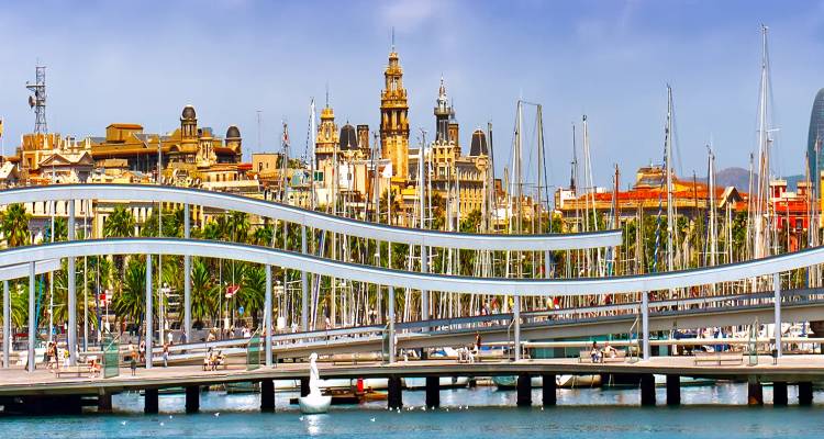 Vue dynamique à travers l'eau vers la ligne d'horizon historique de Barcelone avec la passerelle ondulante Rambla del Mar et les yachts de la marina sous un ciel bleu dégagé.