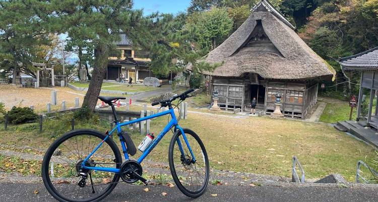 Bicicleta de carretera azul estacionada frente a un templo rústico de madera con techo de paja en medio de terrenos boscosos en el Japón rural.