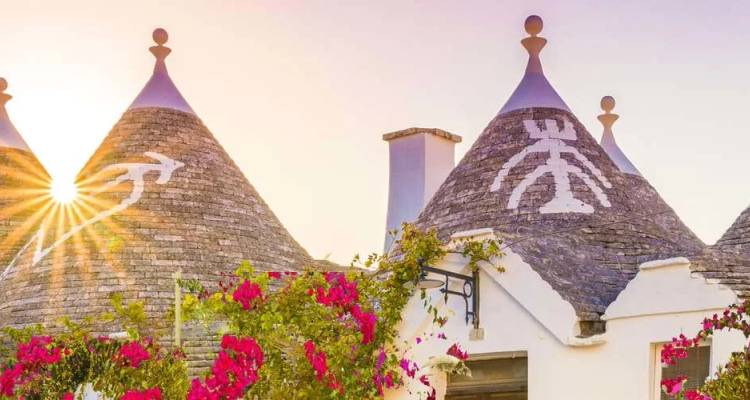 Les maisons trulli iconiques aux toits de pierre coniques décorés de symboles blancs, encadrées par des bougainvillées rose vif et un coucher de soleil.