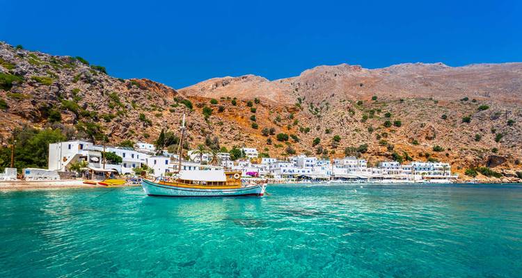Ein kleines Fischerboot schwimmt auf kristallklarem, türkisfarbenem Wasser vor dem weiß getünchten Küstendorf, das von zerklüfteten Hügeln umgeben ist.