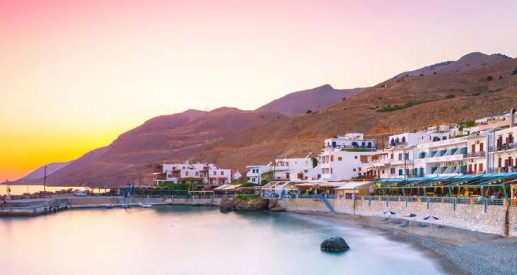 Whitewashed seaside houses line a tranquil bay beneath pastel sunset skies and rugged hills.